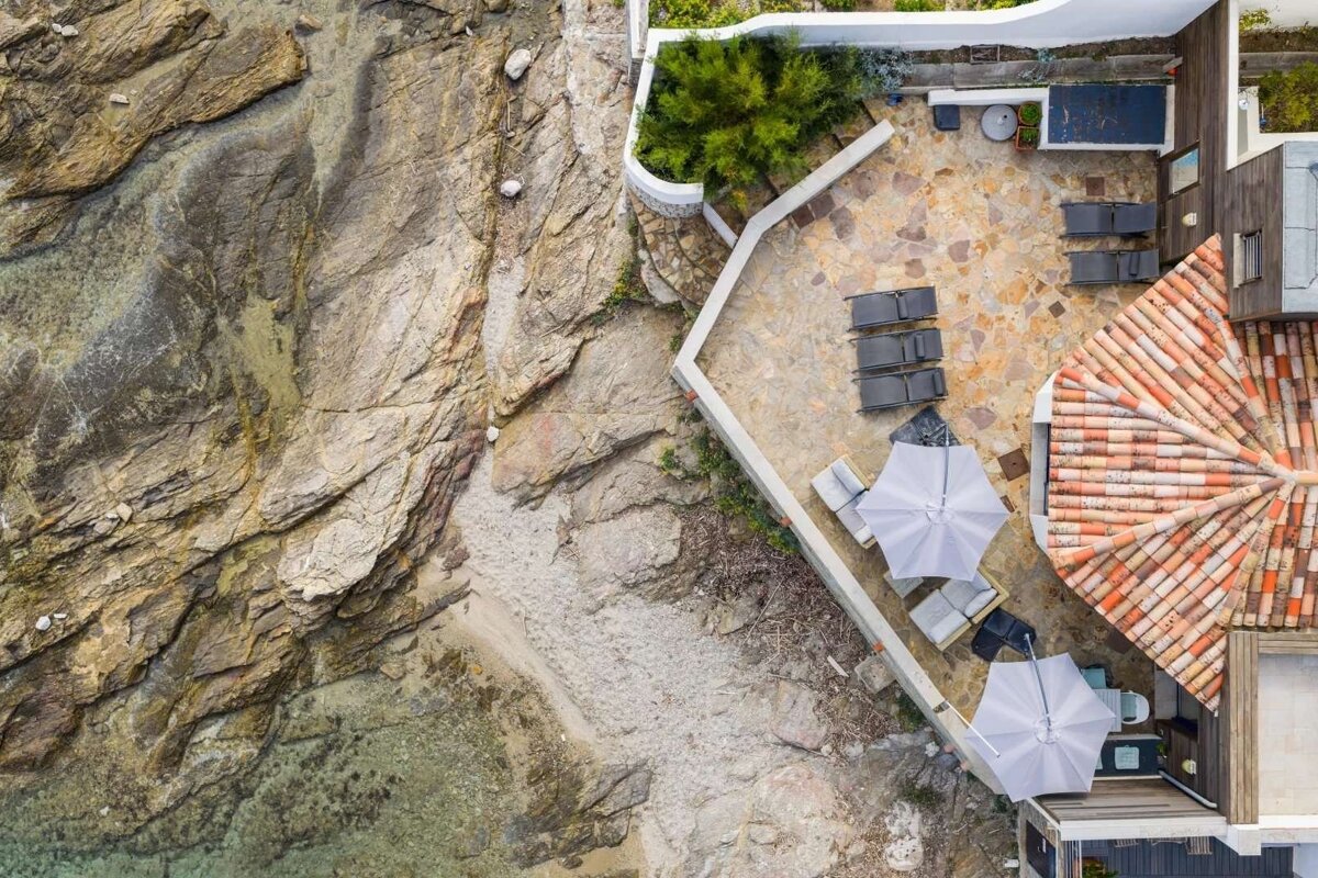 Aerial view of a villa's patio and tiled roof next to a rocky beach and clear water, featuring outdoor furniture and umbrellas.