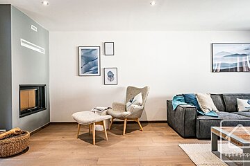 A modern living room features a built-in fireplace on a gray wall, a light armchair with ottoman, a dark sofa, and abstract blue artwork on white walls, all on a wooden floor.