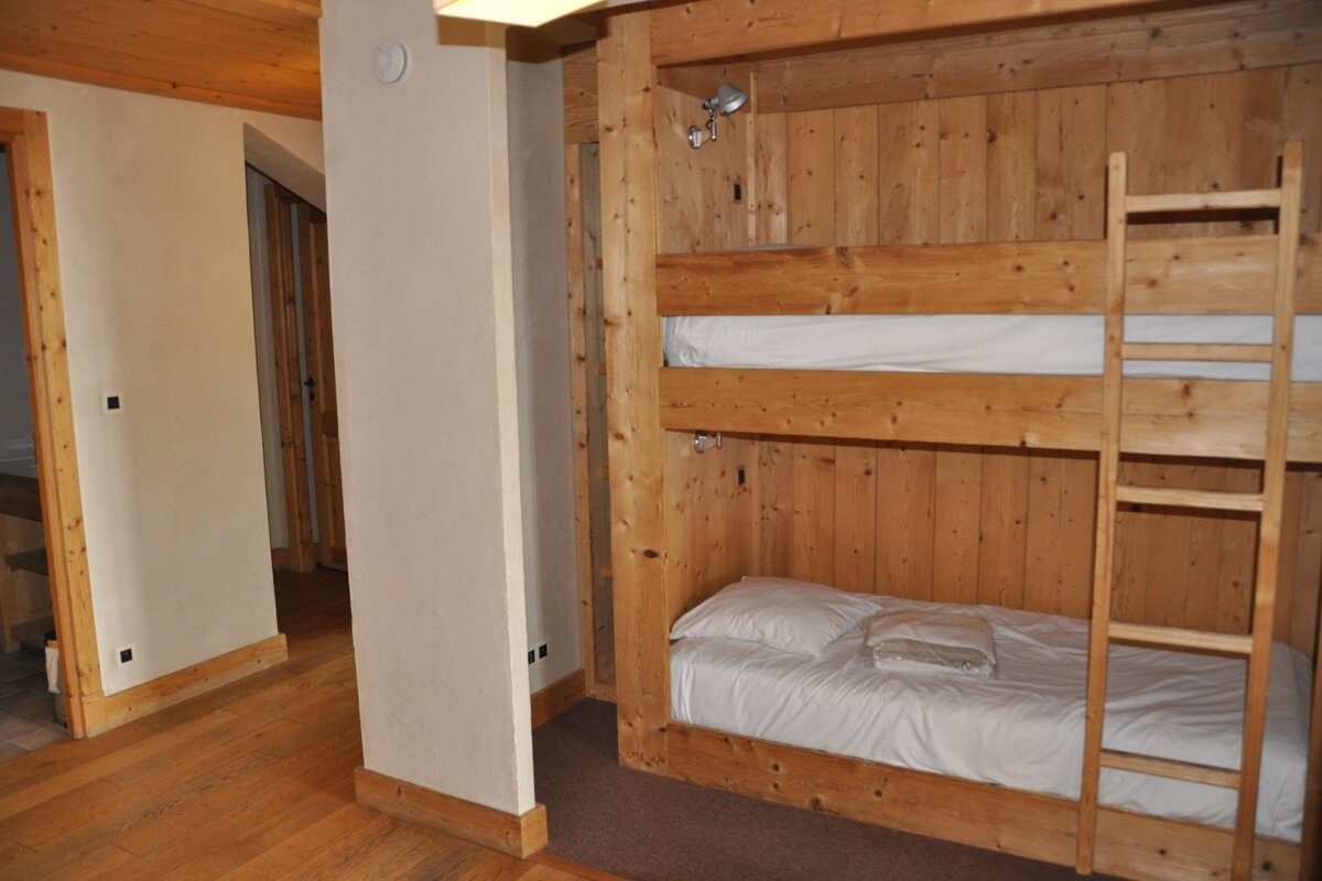 A cozy, rustic room features wooden bunk beds with white bedding built into a wood-paneled wall. A ladder provides access to the top bunk. Wooden flooring extends into a hallway.