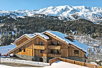 A wooden chalet with snow on the roof is surrounded by snow covered trees