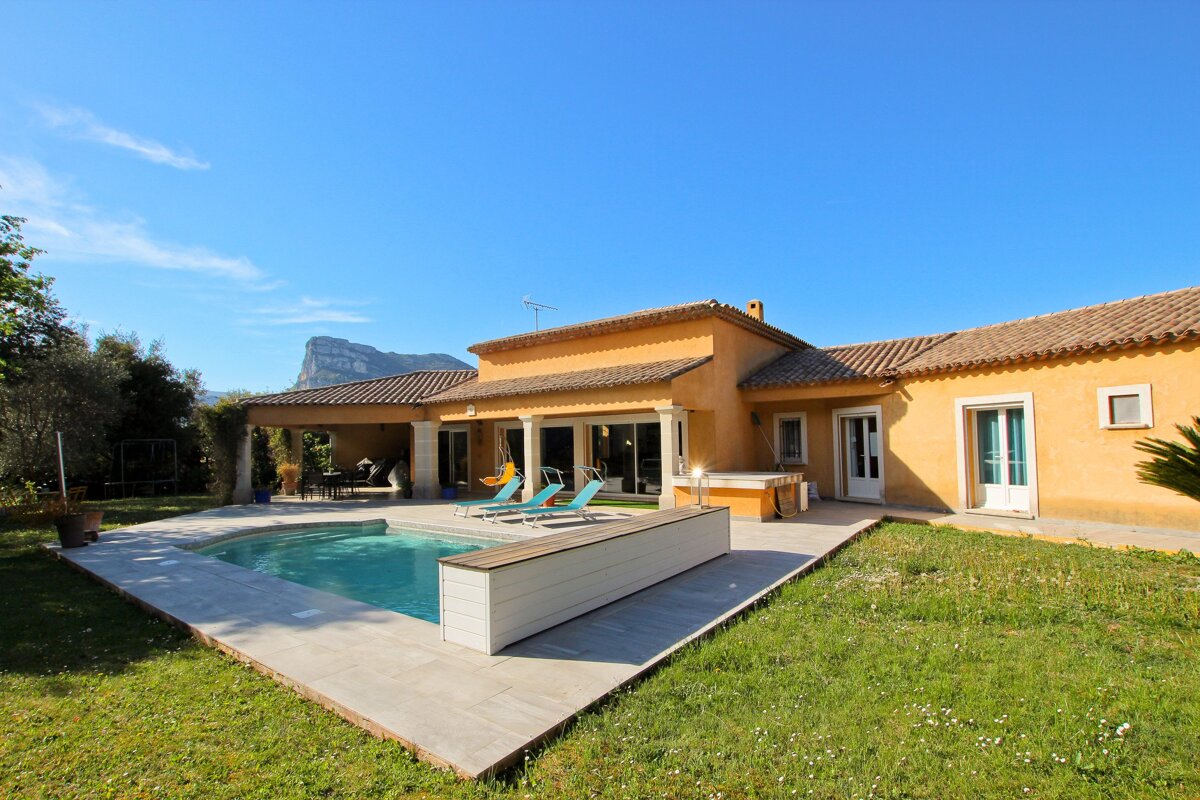 A sunny, orange-yellow villa with a swimming pool and green lawn, set against a clear blue sky and distant mountains.