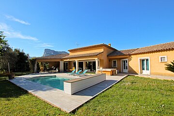 A sunny, orange-yellow villa with a swimming pool and green lawn, set against a clear blue sky and distant mountains.