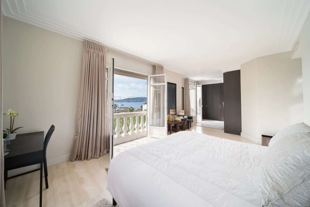 Bright, modern bedroom with a balcony offering a beautiful sea and bay view. Features white bedding, light walls, and a small desk area.