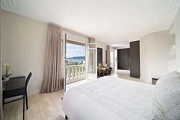 Spacious bedroom with a balcony overlooking a bay, featuring a white bed, desk, and dark built-in wardrobes in a modern, bright setting.