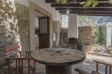A rustic outdoor patio with stone walls, a wooden spool table, striped chairs, and an open doorway. Vines hang from a beamed ceiling.