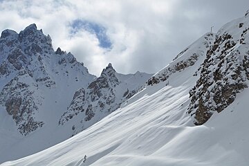 Snowy, jagged mountain peaks rise under a cloudy winter sky, with a steep snow-covered slope in the foreground.
