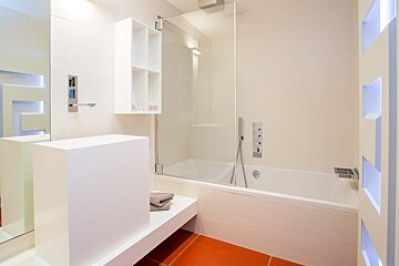 A minimalist white bathroom featuring a bathtub with a glass partition and a vibrant orange tiled floor.