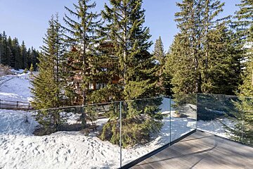 A modern glass balcony with a wooden floor overlooks a snowy forest scene with tall pine trees and a traditional wooden chalet.