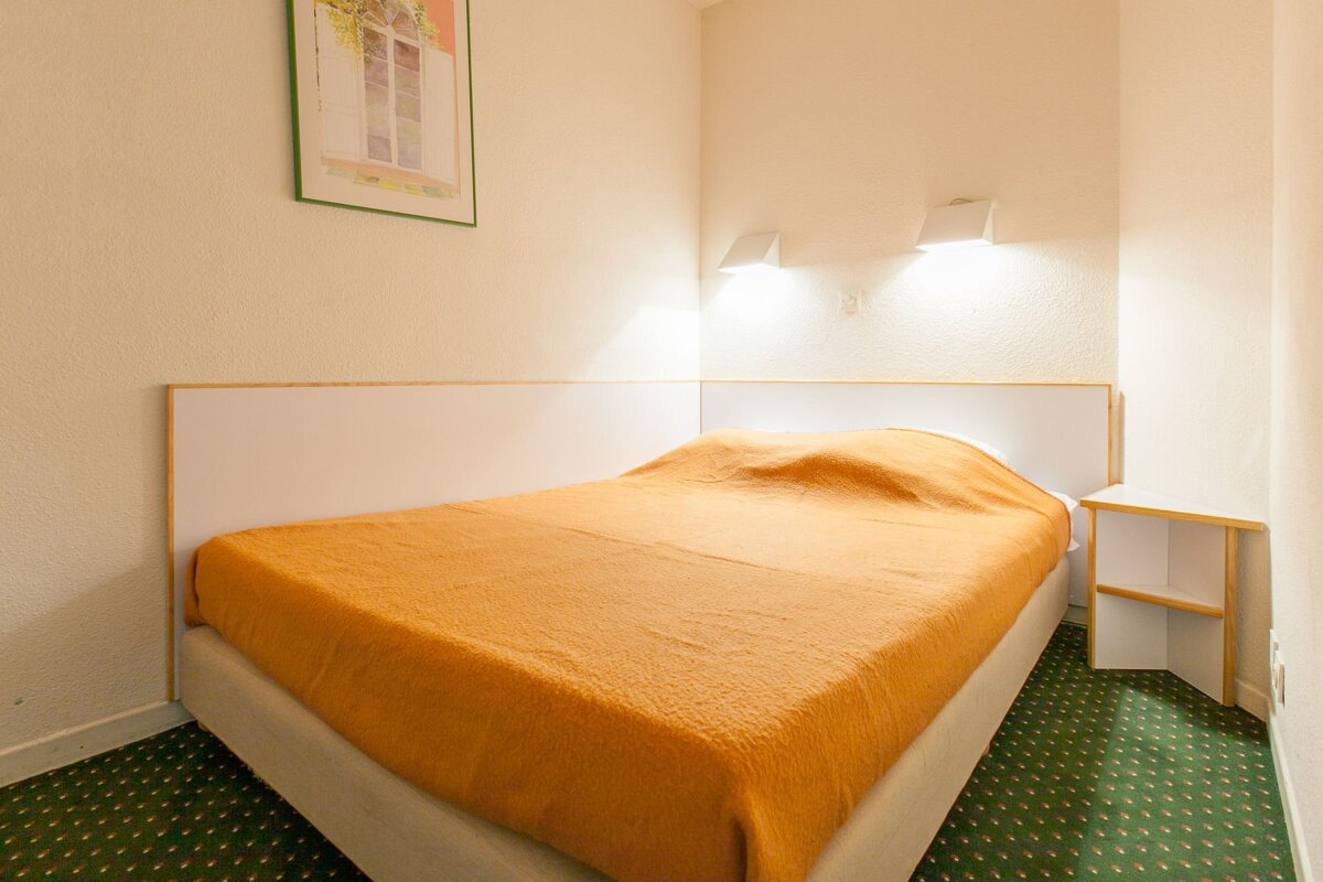 A functional bedroom with an orange blanket on the bed, white headboard, white walls, green carpet, and wall lights.