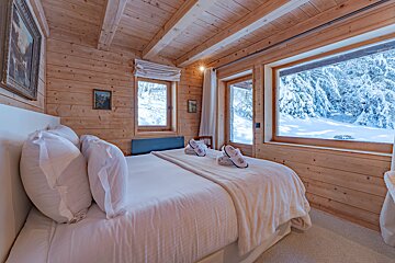 Cozy wooden cabin bedroom with a plush white bed and large windows overlooking a beautiful snow-covered winter forest.