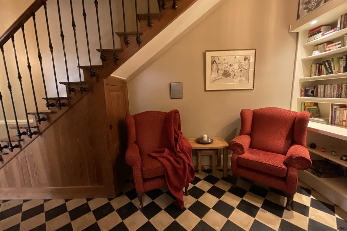 Two red armchairs on a checkered floor sit under a wooden staircase, flanked by a bookshelf, creating a cozy reading nook.