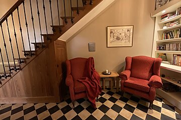 Two red armchairs on a checkered floor sit under a wooden staircase, flanked by a bookshelf, creating a cozy reading nook.