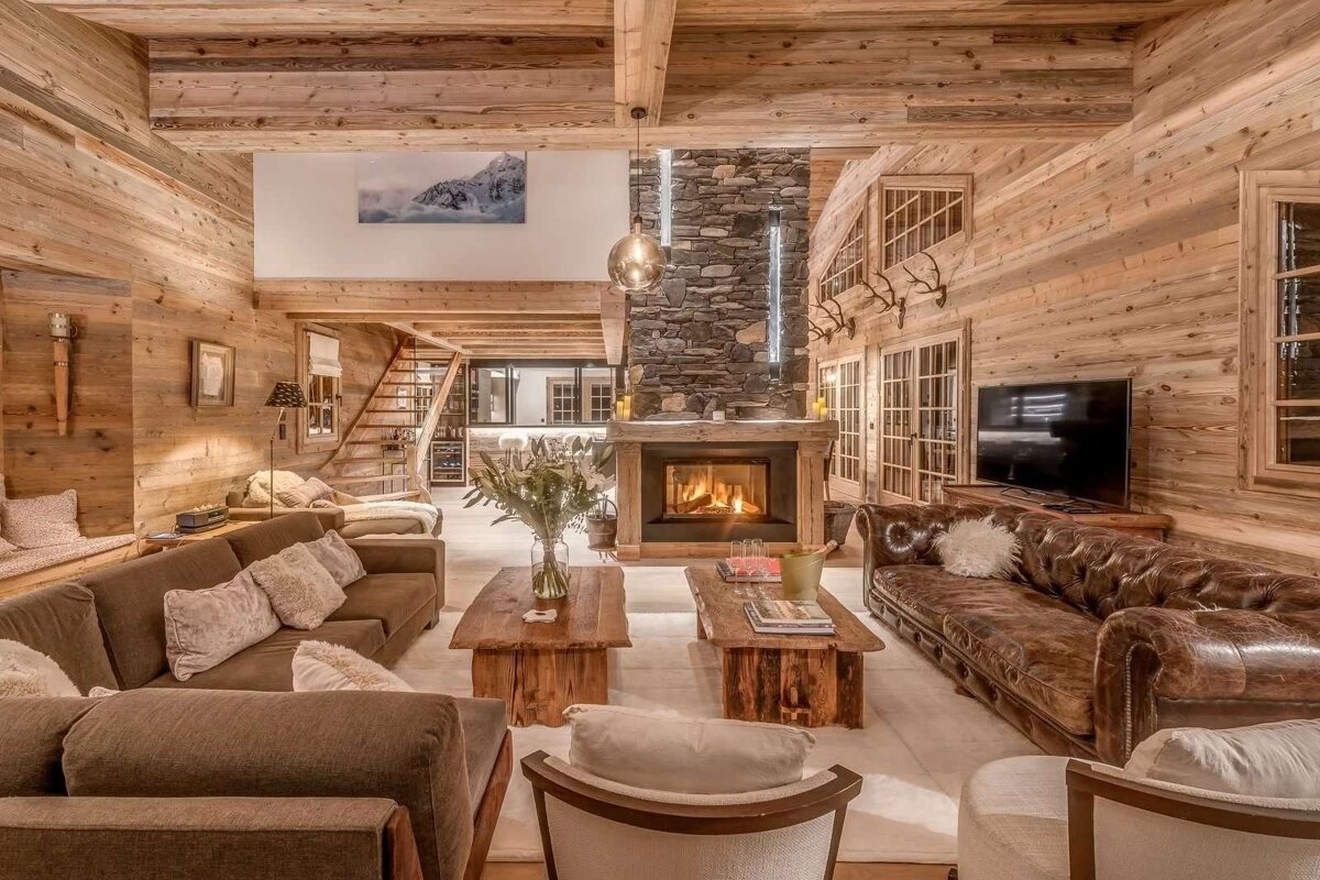 Luxurious chalet living room featuring rustic wood paneling, a grand stone fireplace, plush sofas, and mountain views for a cozy, upscale ambiance.