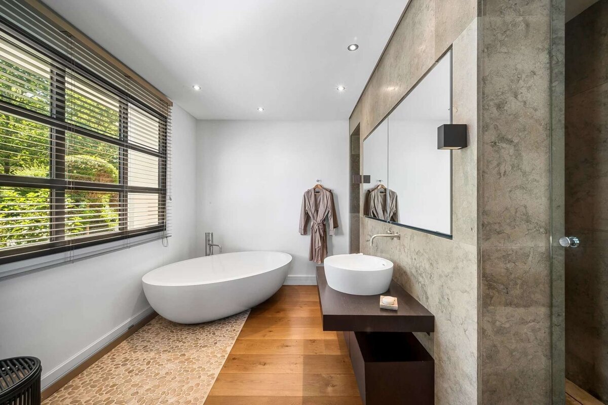 Stylish modern bathroom with a freestanding tub on a pebble path, wood floors, large window, sleek vanity, and a cozy robe hanging.