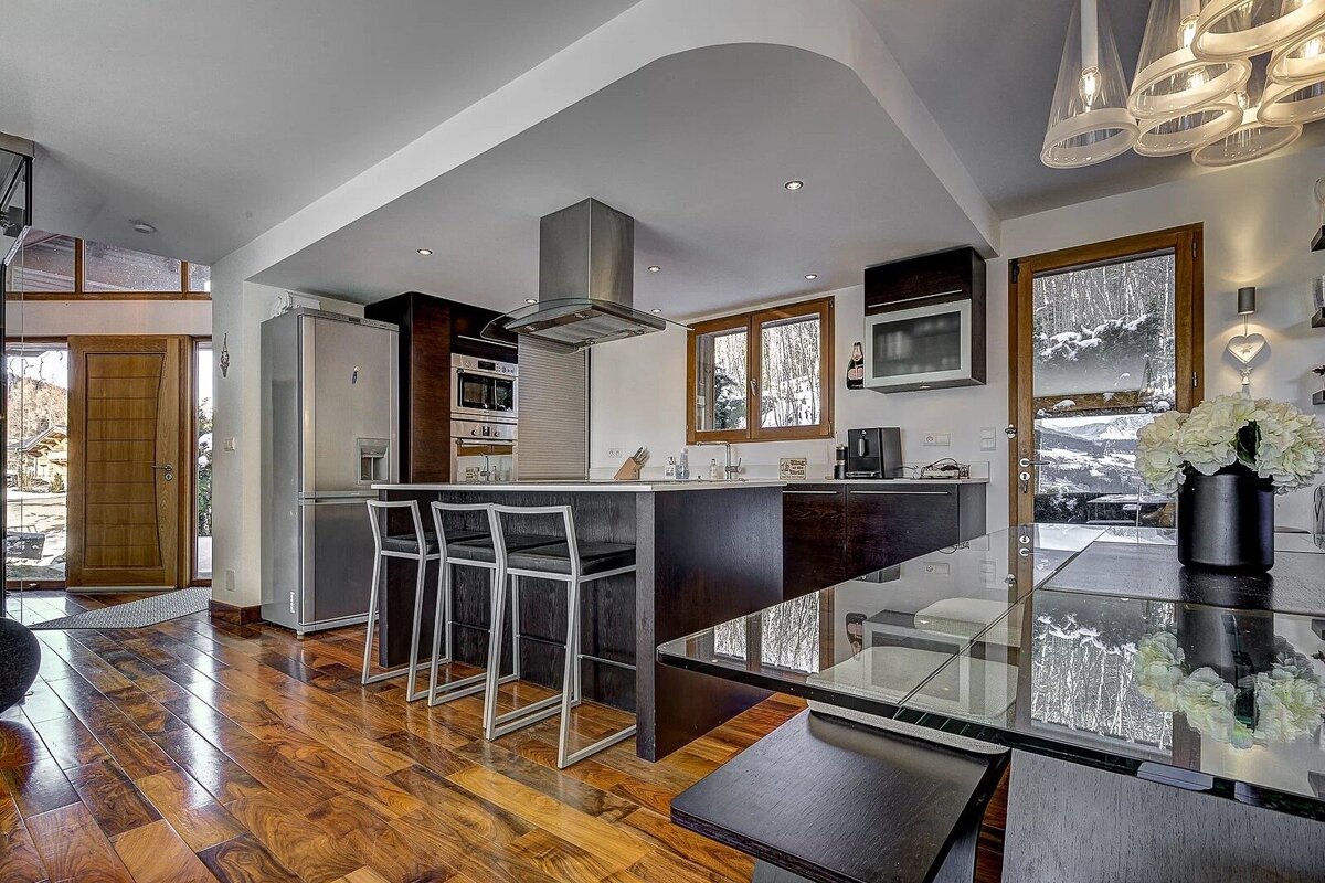 A modern kitchen and dining space with rich wood floors, dark cabinets, an island bar, and large windows overlooking a snowy mountain view.