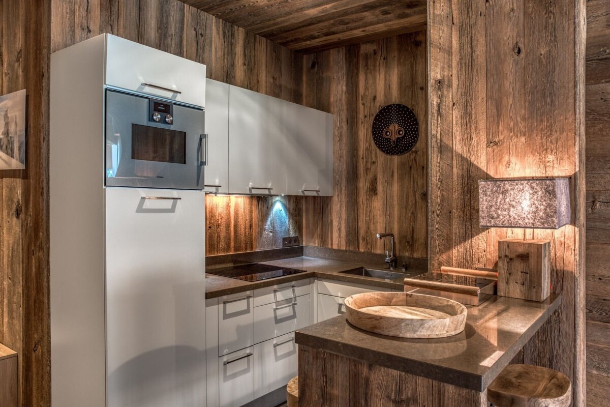 Modern kitchenette in a rustic wooden chalet. Features white cabinets, built-in oven, sink, cooktop, and a lit counter with a lamp and wooden tray.