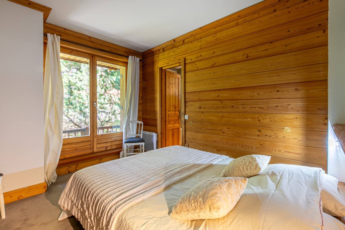 A cozy, chalet-style bedroom featuring warm wood-paneled walls, a large window with green tree views, and a neatly made bed with white linens.