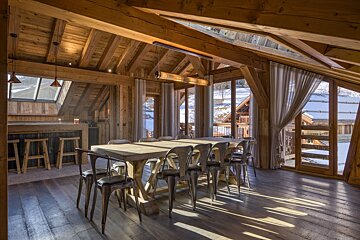 Rustic wooden chalet dining room featuring a long wooden table, metal chairs, and large windows overlooking a snowy mountain landscape.
