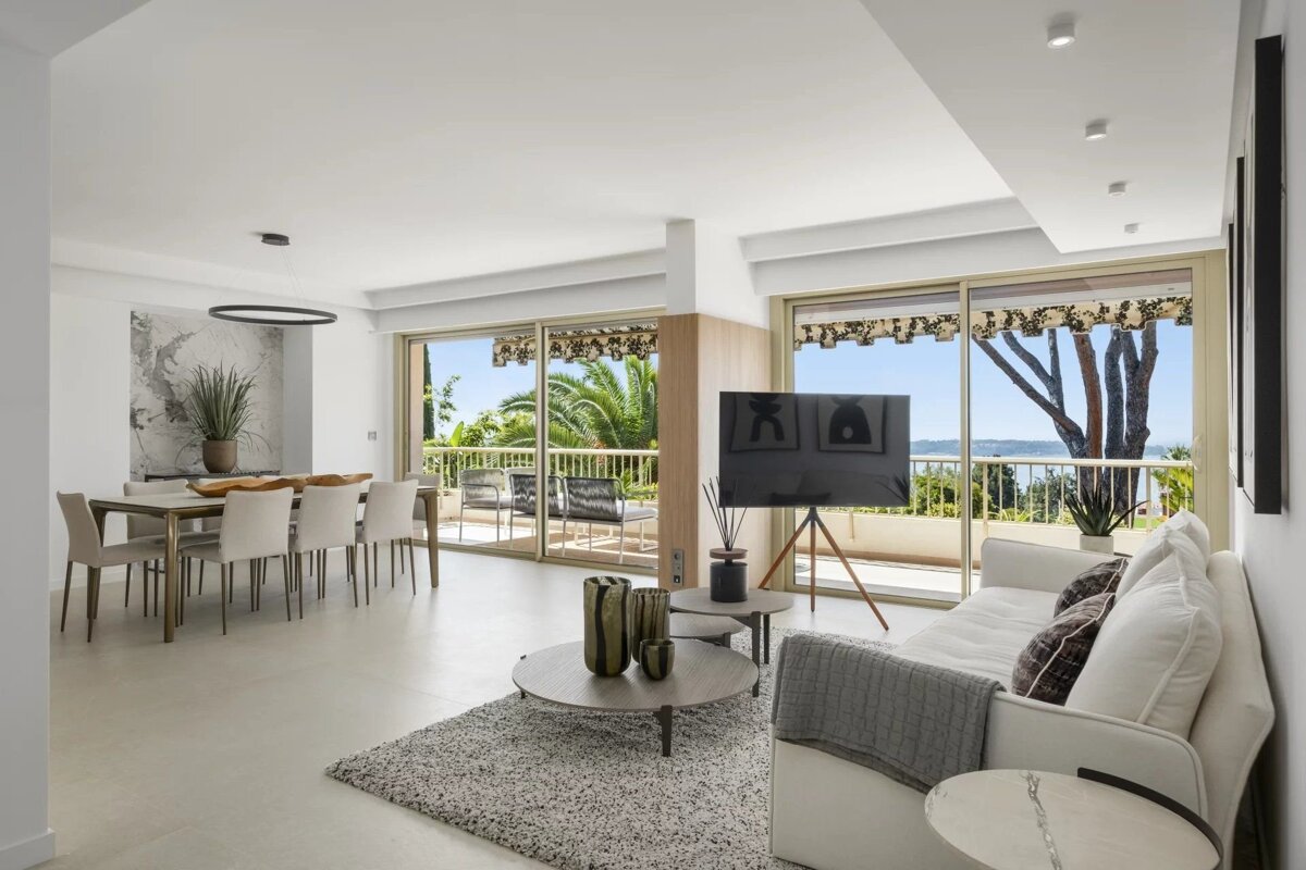 Bright, modern living and dining room with white furniture, a TV, and a balcony overlooking the sea and palm trees.