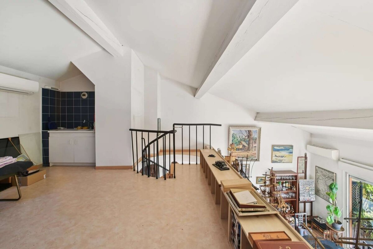 A bright, eclectic attic loft with sloped, beamed ceilings. It features a spiral staircase, a small kitchen nook, and various artwork and collectibles.