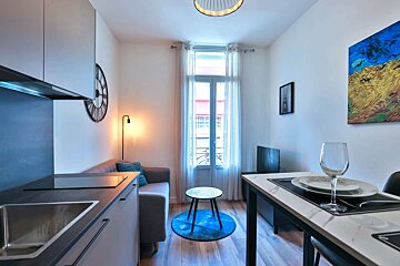 A bright, modern studio apartment features a compact kitchen, living area with a sofa, and a dining space, all on hardwood floors.