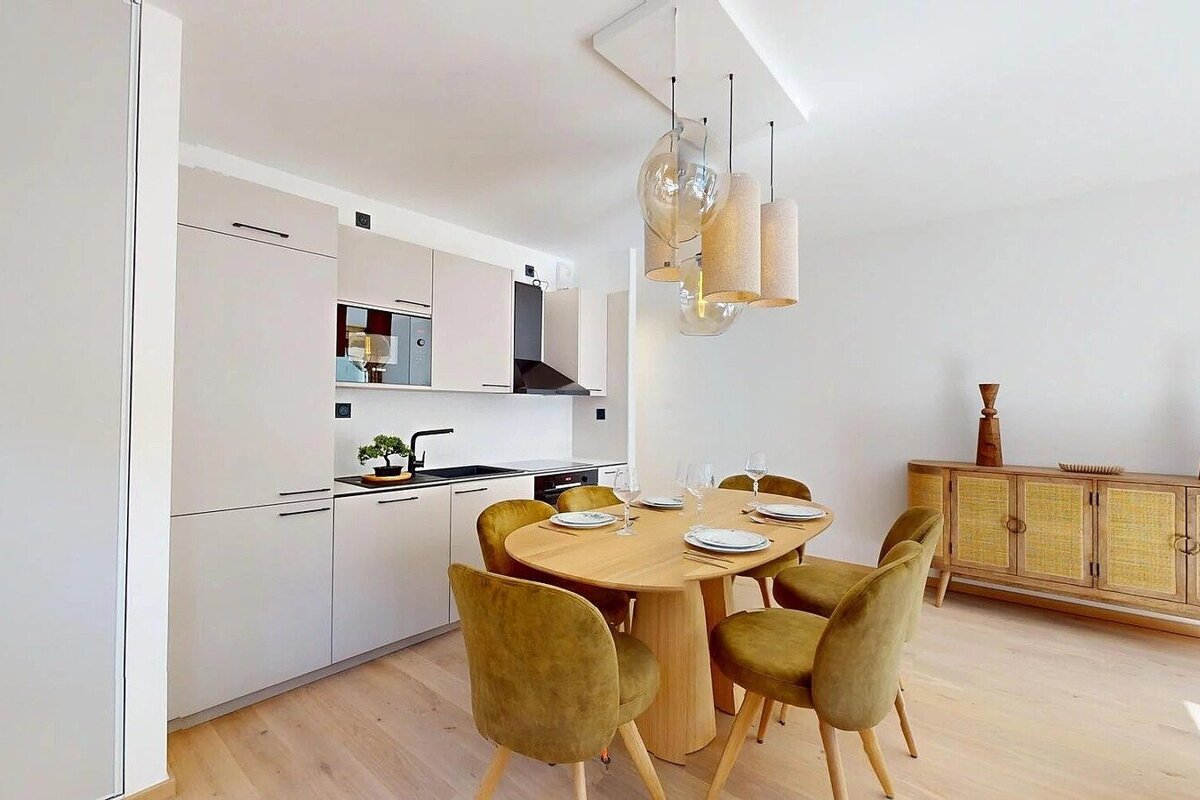 A modern, open-plan kitchen and dining area featuring light wood, off-white cabinetry, olive velvet chairs, and a rattan sideboard.