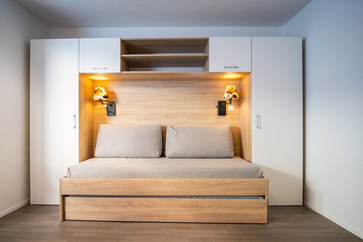 A modern pull-out daybed is integrated into a sleek white and wood wall unit with storage, featuring illuminated wall lamps.