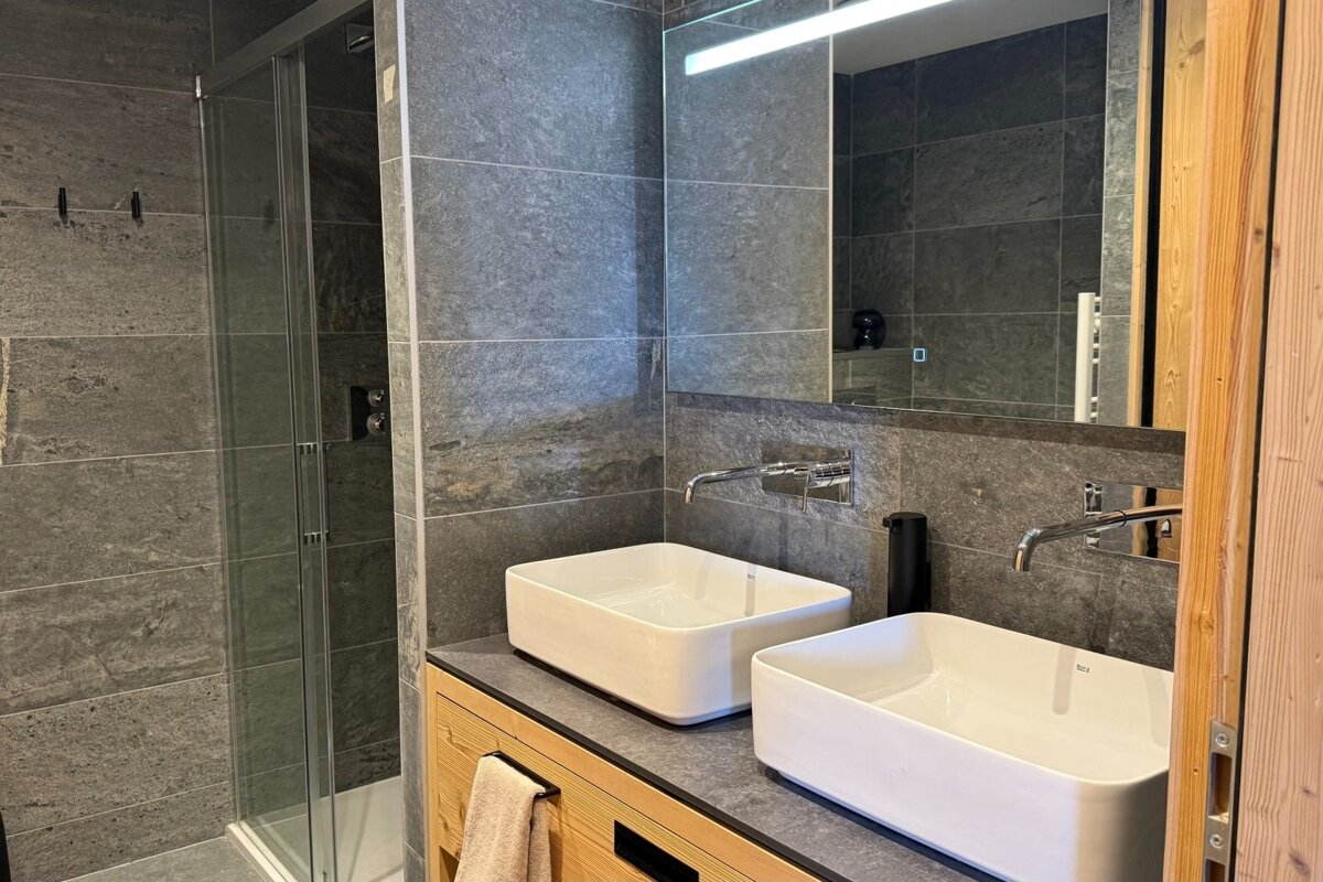 Modern bathroom with grey stone tiling, two white vessel sinks on a wooden vanity, a large mirror, and a glass walk-in shower.