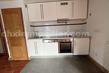 A modern kitchen with white cabinets, stainless steel oven, sink, and dishwasher. Features a white tiled backsplash, gray floor, and a wooden door on the left.