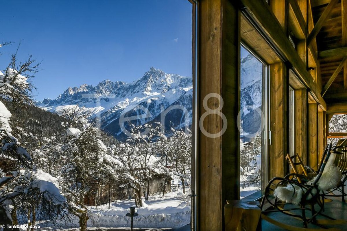 A cozy wooden room with large windows overlooks a majestic snow-capped mountain range, snow-covered trees, and a bright blue sky on a clear winter day.