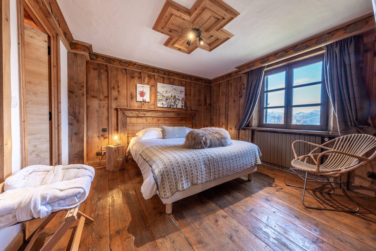 A cozy, wood-paneled chalet bedroom with a large bed, faux fur pillow, mountain view window, and rattan chair, exuding rustic warmth.