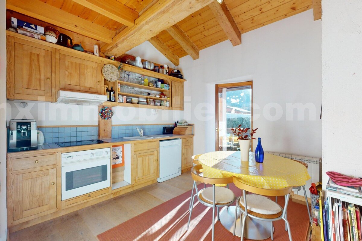 A cozy kitchen features light wooden cabinets, exposed ceiling beams, blue tiled backsplash, and a round dining table with a yellow cloth. A door leads outside.