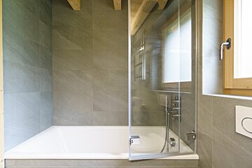 A modern bathroom featuring a white bathtub with a glass shower door, grey tiled walls, and rustic wooden ceiling beams.