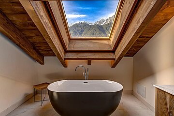 Modern bathroom with a freestanding tub under a skylight revealing snowy mountains and blue sky, framed by rustic wooden beams.