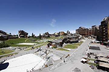 A sunny mountain resort village in summer, with chalet buildings, green hills, a volleyball court, and a white artificial slope under a clear blue sky.