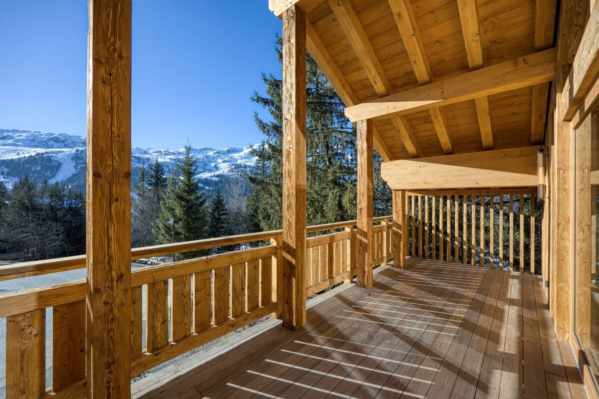 Sunny wooden chalet balcony with panoramic views of snowy mountains, evergreen trees, and a bright blue sky.