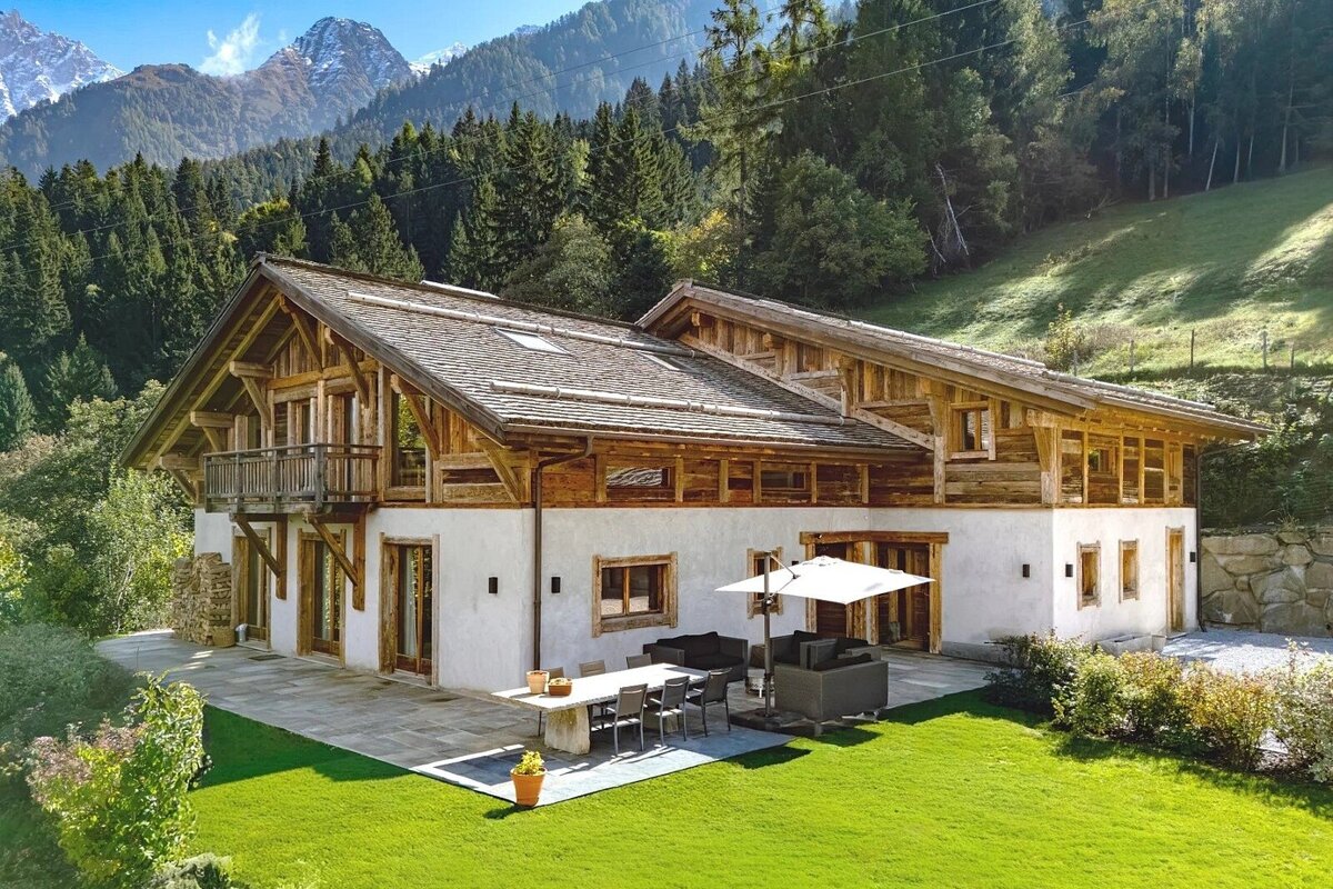 Stunning mountain chalet with a large patio, lush green lawn, and forest backdrop. Snow-capped peaks visible under a clear sky.