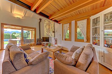 A cozy living room with large sofas, a wood-beamed ceiling, and a fireplace, featuring a sliding door view to a pool and mountains.