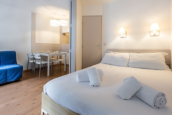 A bright, modern studio apartment with a neatly made bed, a small dining area with a white table, and a blue sofa bed on light wood flooring.