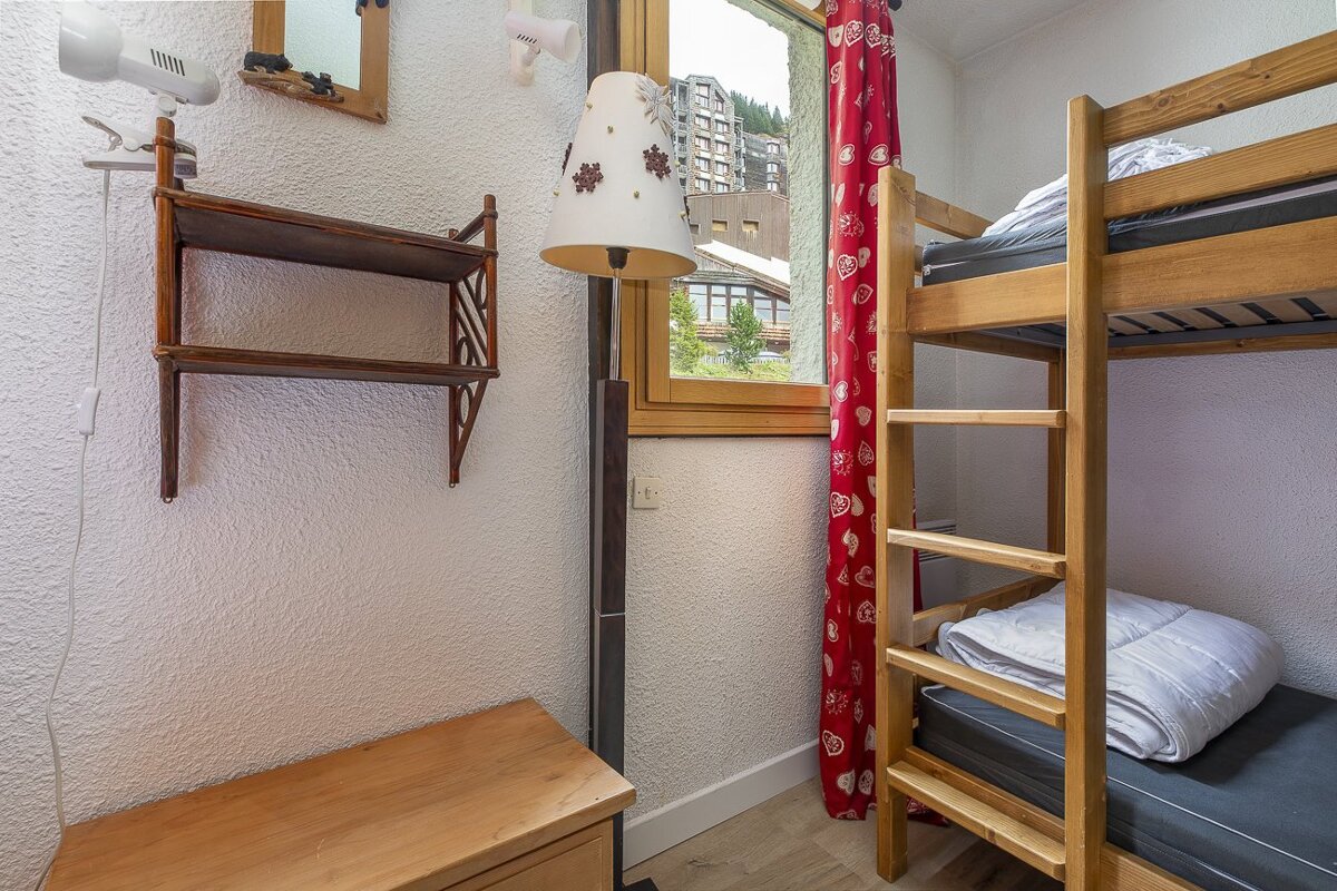 A cozy room with wooden bunk beds, a window with red heart-patterned curtains overlooking buildings, a floor lamp, and wall shelves.