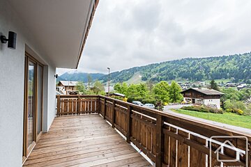 A spacious wooden balcony offers a panoramic view of a lush green mountain village, forested slopes, and cloudy sky.