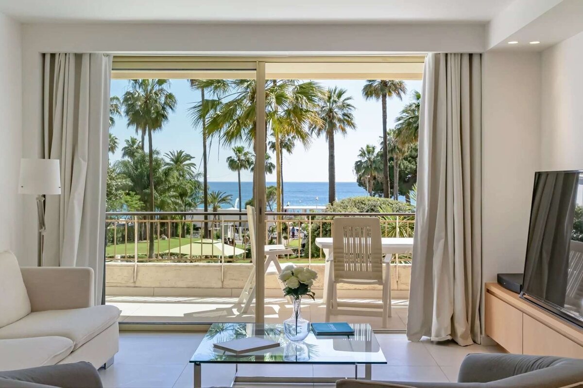 A bright living room with a glass door balcony overlooking palm trees, a green lawn, and the sparkling blue ocean.
