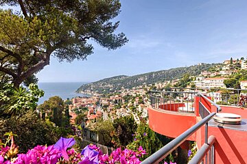 Scenic coastal town on hills overlooking a bay, with a red balcony and vibrant purple flowers framing the view.