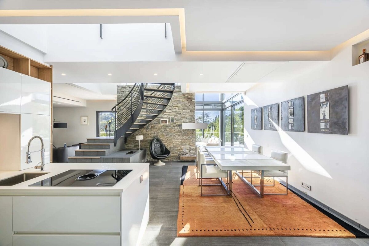 Modern open-plan space featuring a sleek kitchen island, a dining area, a spiral staircase by a stone wall, and large sunlit windows.
