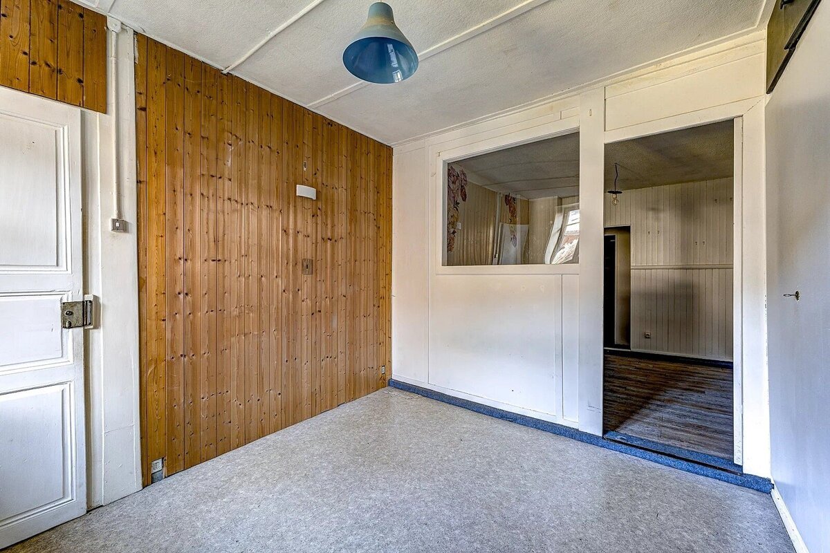 An empty room with wood-paneled wall, white door, blue ceiling lamp, and a framed opening to an adjacent room. Light grey floor.