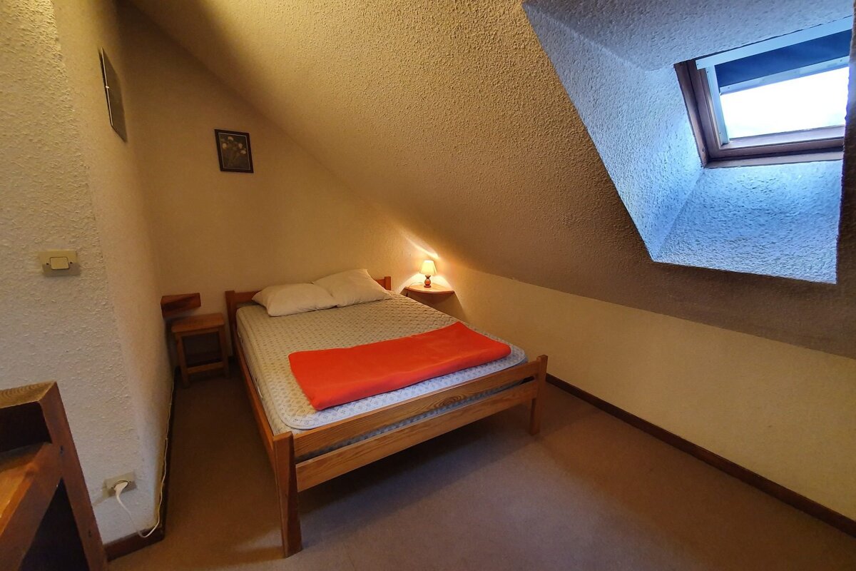 A cozy attic bedroom with a single bed, orange blanket, sloped ceiling, and a bright skylight window. Features warm ambient lighting.