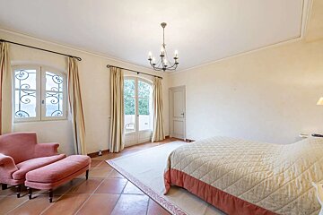 A spacious bedroom features a quilted bed, pink armchair with ottoman, terracotta floor, arched windows with curtains, and a central chandelier.