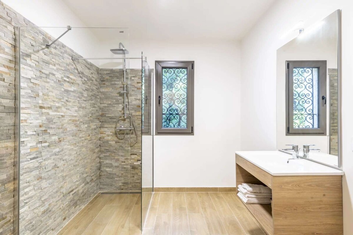 Modern bathroom featuring a stone-tiled walk-in shower, wooden vanity, light wood floor, and a window with decorative metal grilles.