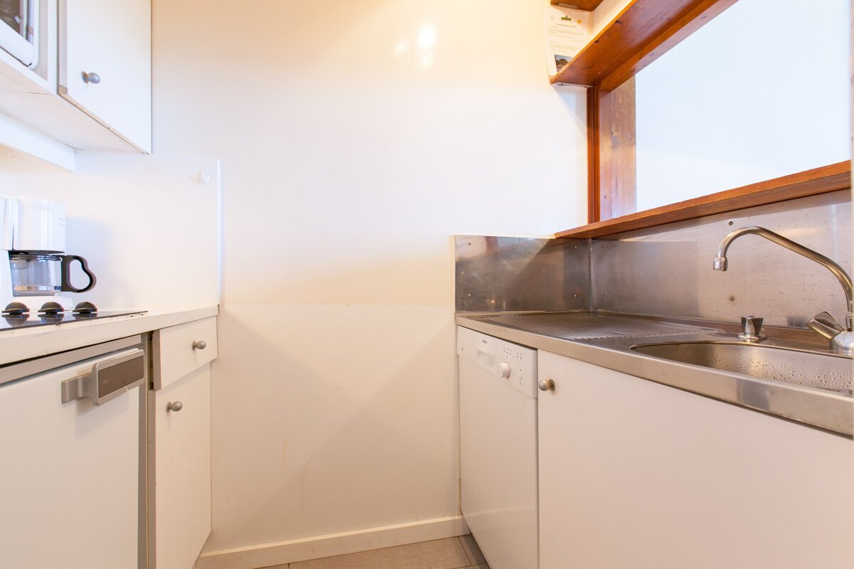 A small, clean kitchen with white cabinets, stainless steel sink and counter, a coffee maker, dishwasher, and a wooden pass-through window.