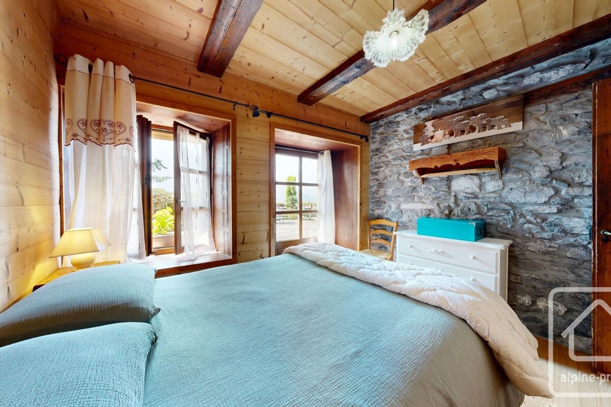 A cozy, rustic bedroom with light wood walls, ceiling, and a stone accent wall. A large bed sits by two open windows offering bright views.